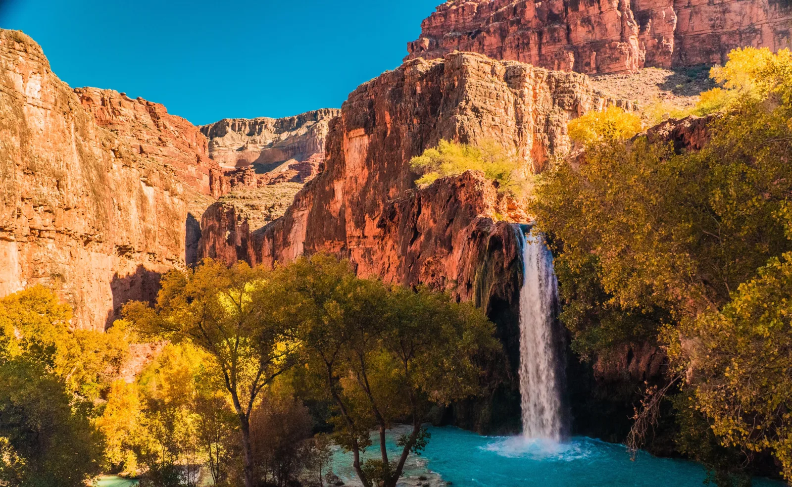 Waterfall dropping into a turquoise pool below red rock and autumn trees